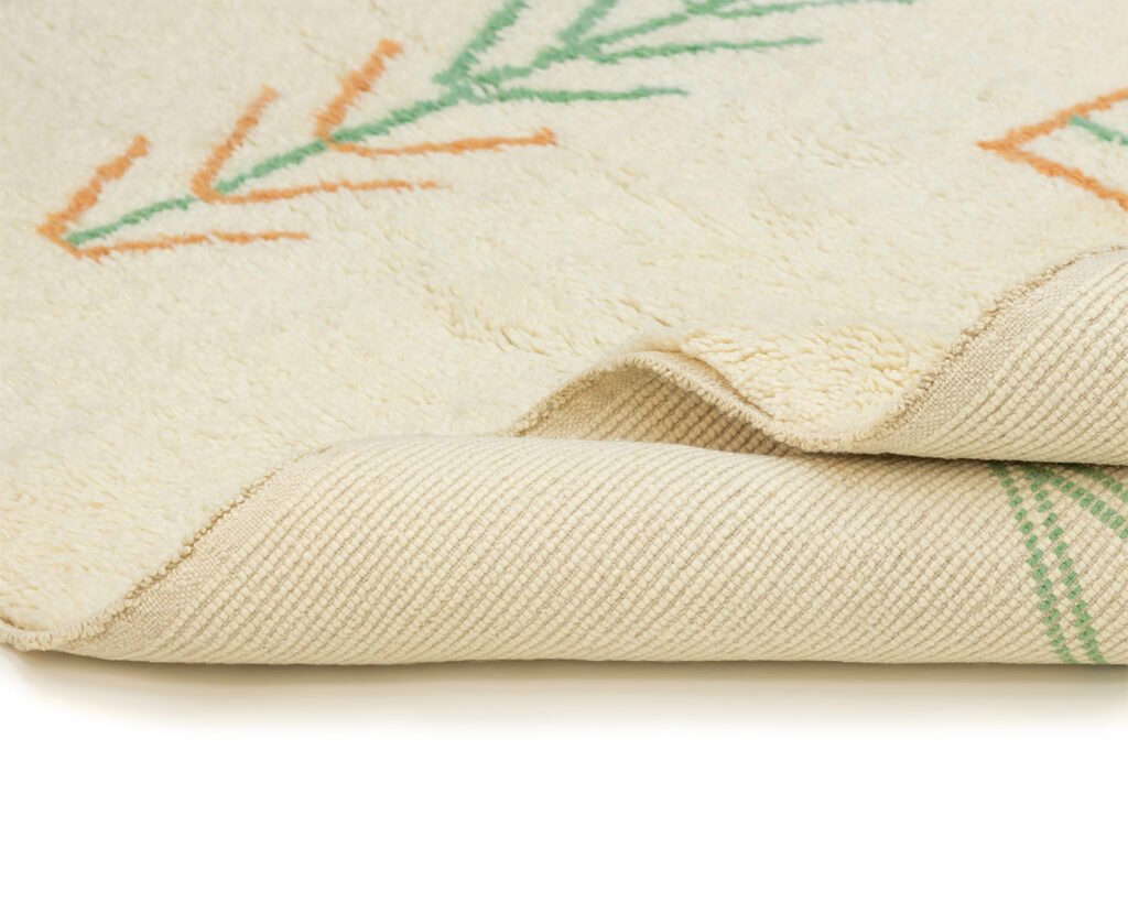 Close-up of the corner of an artisanal rug, showcasing the beige fabric adorned with abstract green and orange patterns. The texture of the hand-knotted design is visible, offering a glimpse of its authentic craftsmanship from Morocco.
