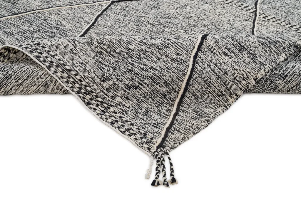 The description unveils a close-up of a black and white patterned rug corner adorned with tassels. Its intricate geometric line designs and rich texture add an elegant touch to any space, making it a pivotal decor piece.
