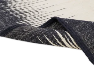 The intricate detail of a woven rug is revealed as its corner folds back, showcasing a striking black and white striped pattern.