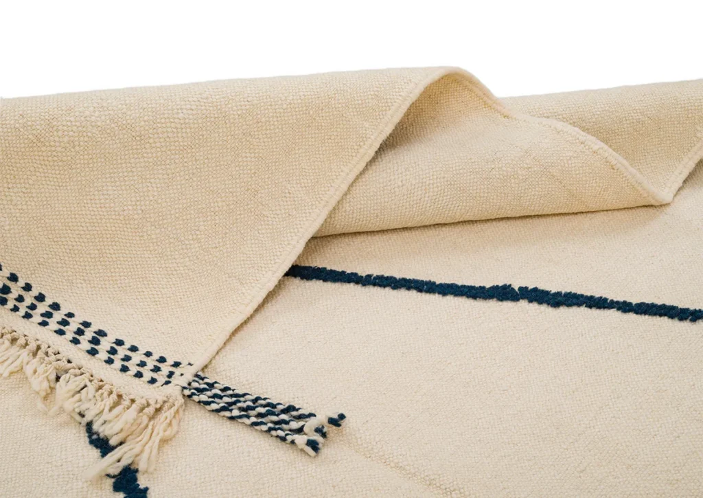 A beige textured flatweave rug with folded edges showcases navy tassel detailing and a diagonal navy line design.