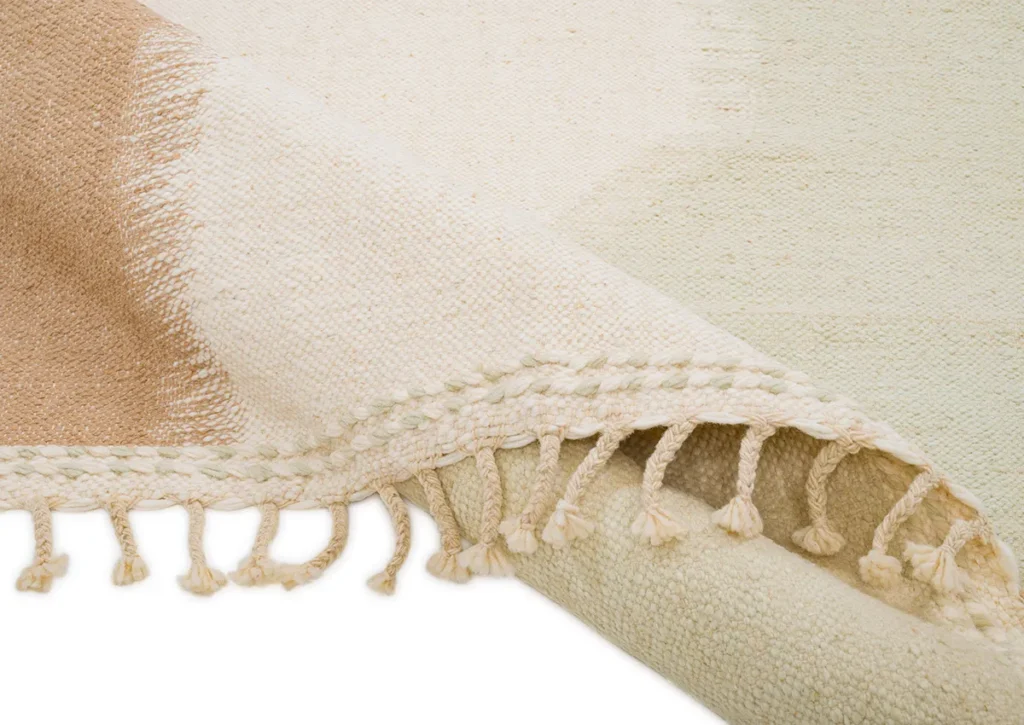 Close-up of a beige and cream flatweave rug with fringe detail along the edge. The fabric is folded at the corner, highlighting its texture and craftsmanship.