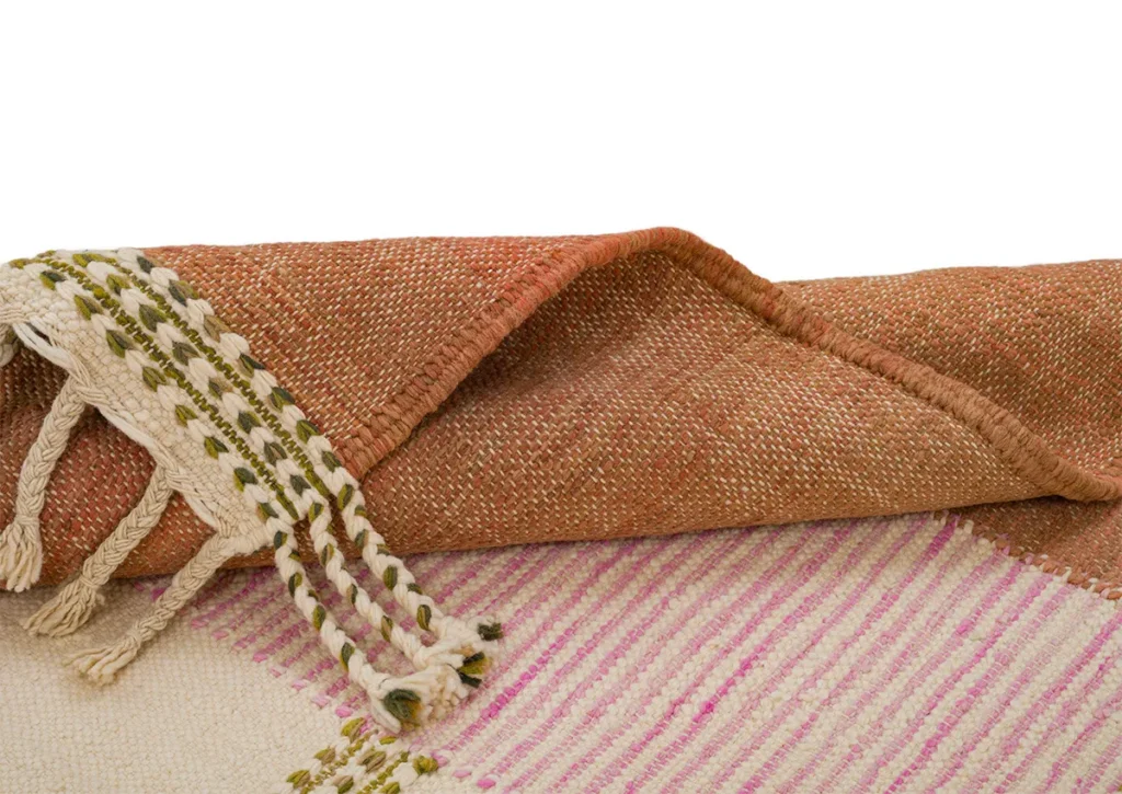 A handwoven rug, featuring pink and red stripes, showcases tasseled edges interlaced with green and brown threads.
