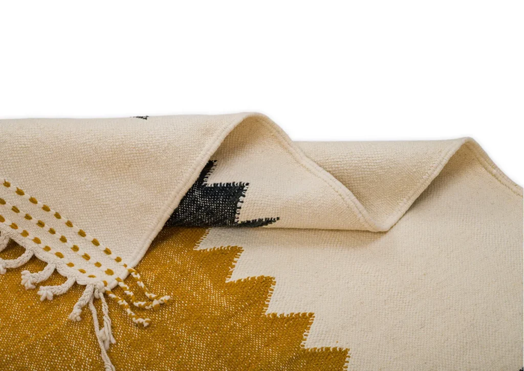 Close-up of a folded woven textile with a zigzag pattern in mustard yellow and black on a cream background, featuring fringed edges. The intricate design adds an artistic touch to any space, making it a striking accent piece.