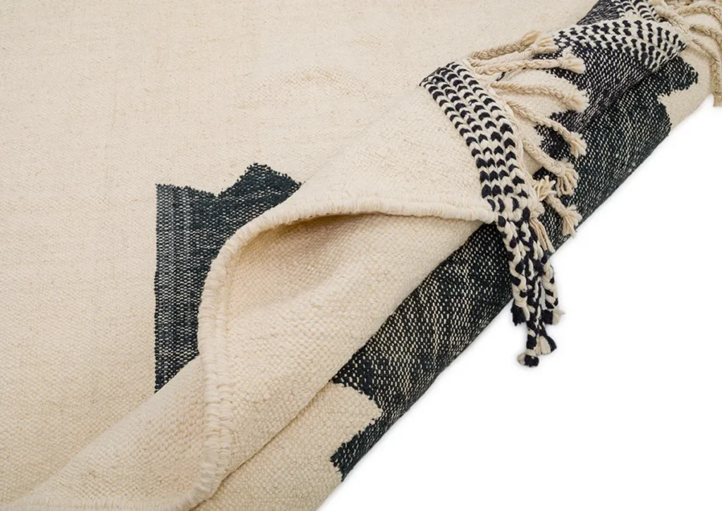 Close-up of a beige handwoven rug with black geometric designs and fringed edges, partially folded over.