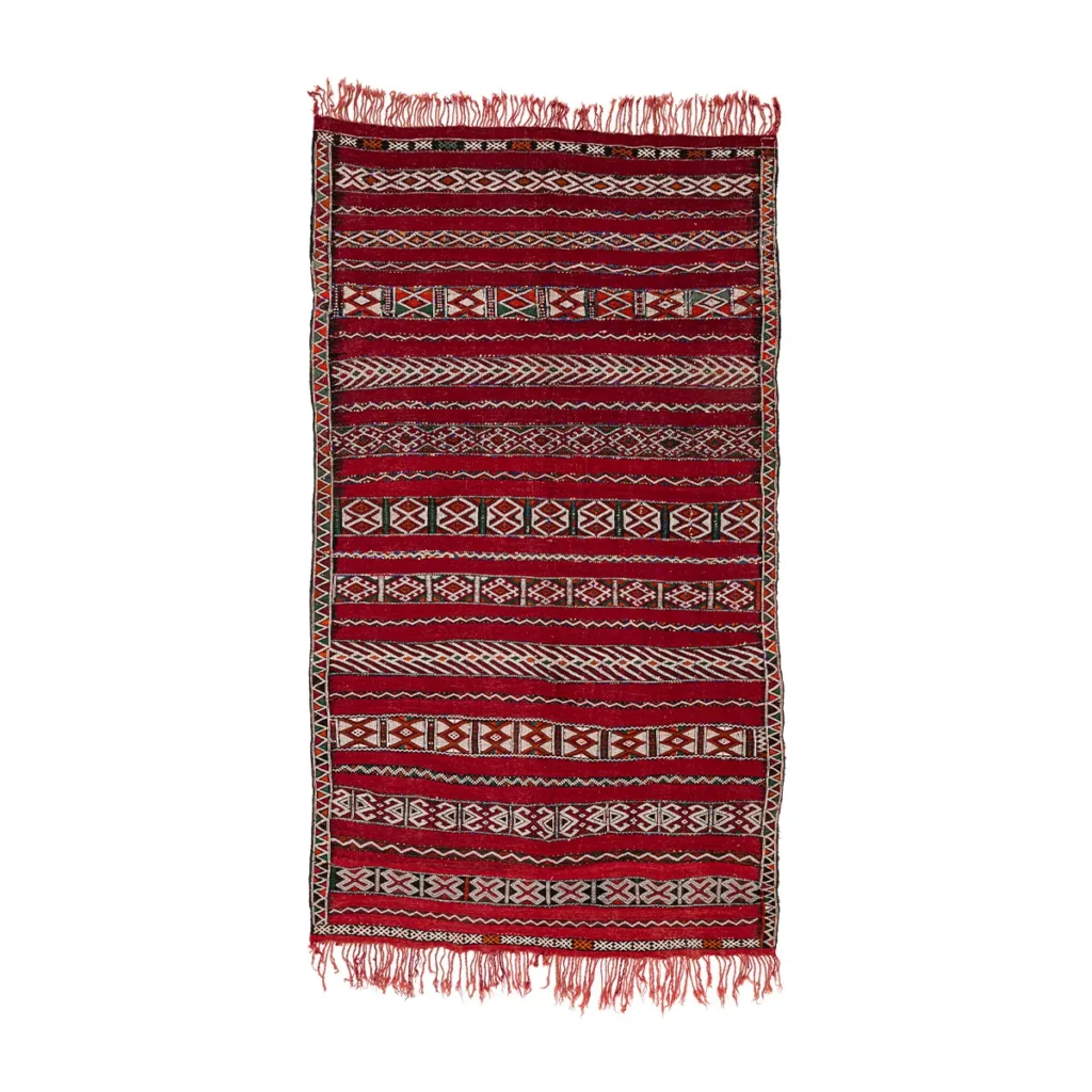 A vintage rectangular red and white patterned flat rug with fringed edges, featuring geometric designs and horizontal stripes.