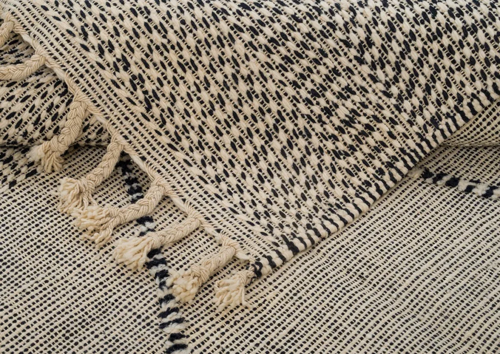 Close-up of a handwoven rug in black and white, featuring tassels. The pattern consists of small, irregular shapes that create a textured appearance.