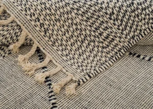 Close-up of a handwoven rug in black and white, featuring tassels. The pattern consists of small, irregular shapes that create a textured appearance.