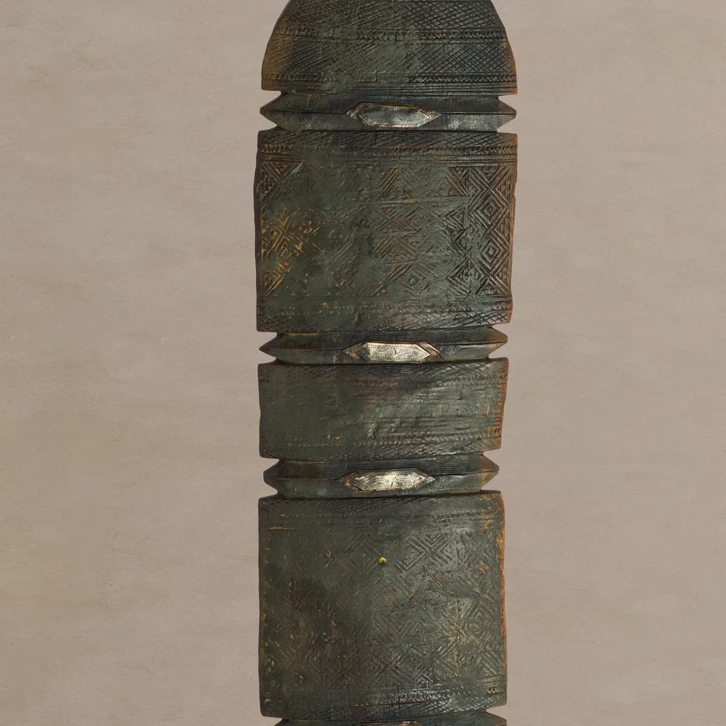 Close-up of a hand-carved, cylindrical object with intricate geometric patterns and metallic accents, reminiscent of a vintage wooden tent peg, set against a plain background.