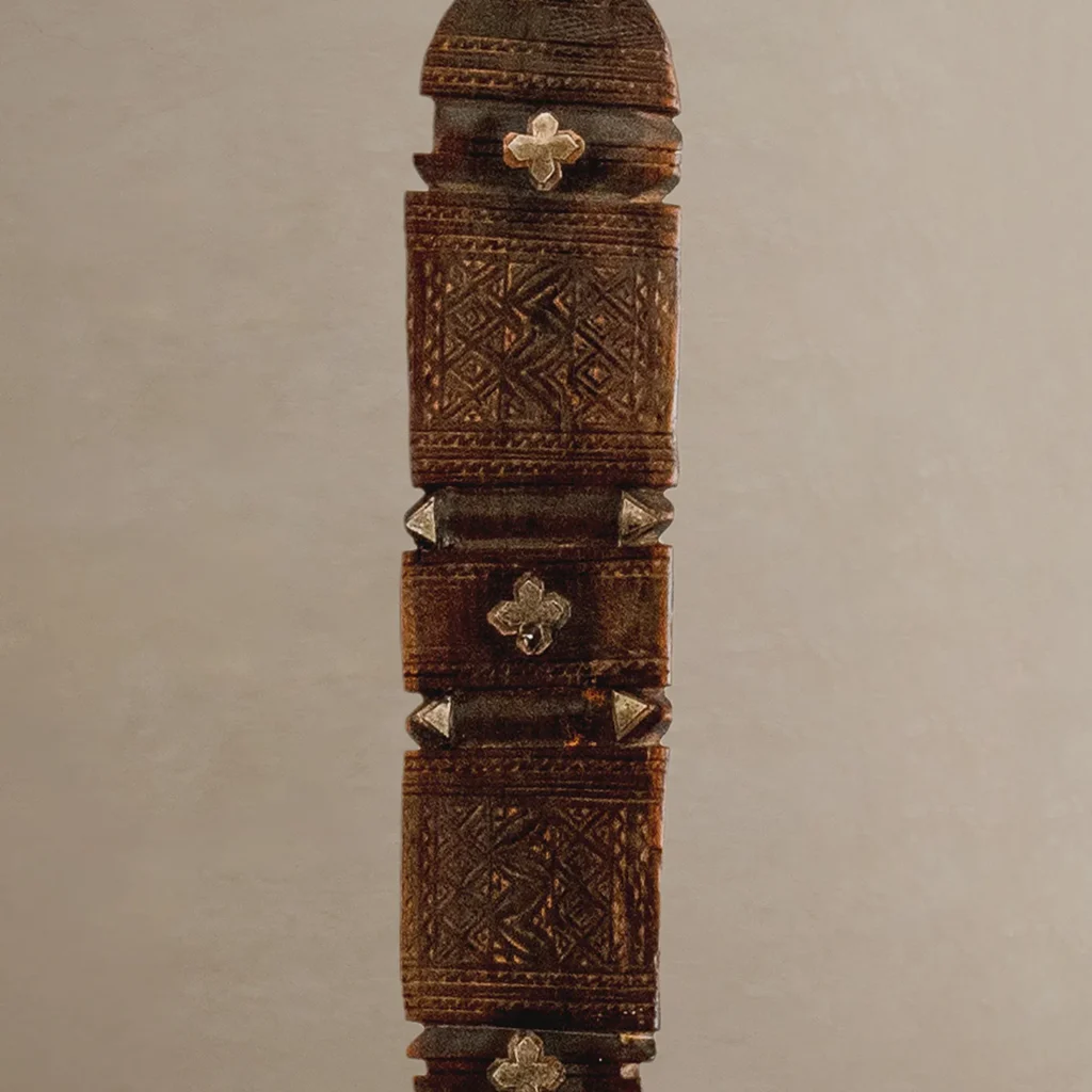 Close-up of a hand-carved wooden stick or staff adorned with intricate geometric carvings and metallic decorative elements, reminiscent of a vintage wooden tent peg.