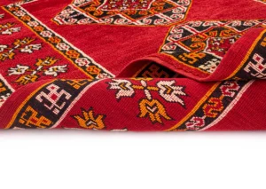 A beautifully crafted red and multicolored woven rug featuring intricate geometric and floral designs, with one edge elegantly rolled.