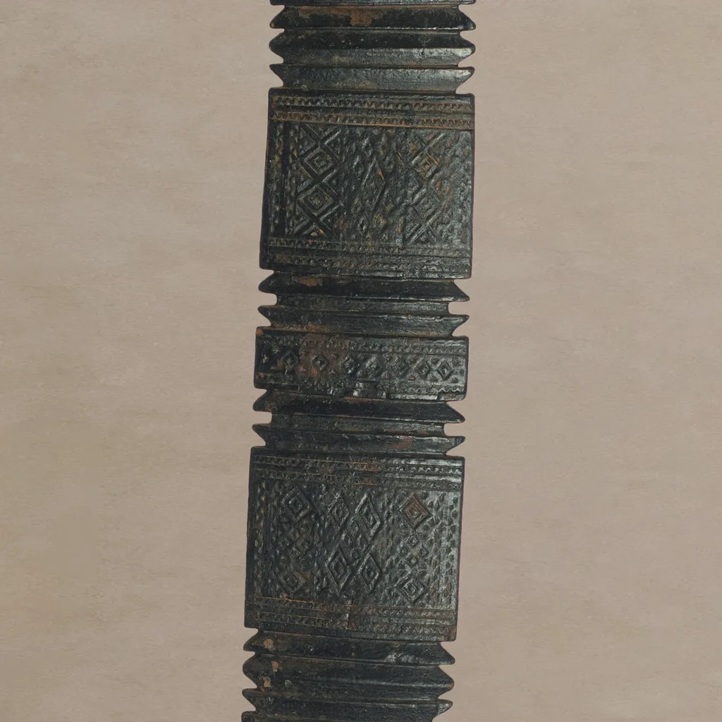 Hand-carved vintage wooden object with intricate geometric patterns and ridges, reminiscent of an ornately designed tent peg, shown against a neutral background.