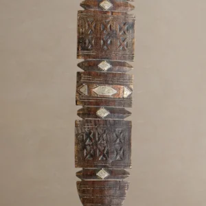 An intricately carved wooden pillar, reminiscent of vintage wooden tent pegs, features geometric and diamond-shaped patterns, displayed on a light brown background.