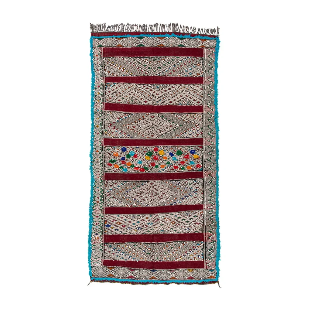 A vibrant woven rug with geometric patterns, showcasing bold red, white, and multicolored sections, framed by a striking blue border.