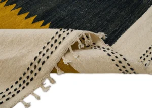 Close-up of a handwoven rug with a folded corner, showcasing a black and mustard zigzag pattern alongside black dashed lines on a cream background, finished with elegant cream tassels.
