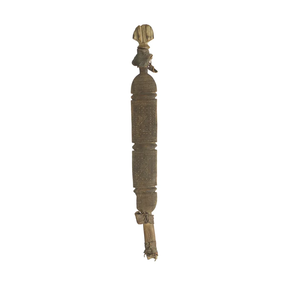 A hand-carved vintage wooden tribal staff with intricate details and a small figure on top. The staff, reminiscent of an ornate tent peg, is adorned with geometric patterns and shows signs of wear.