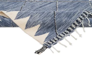A partially folded blue and white handwoven rug with a zigzag design and fringe detailing on the ends.