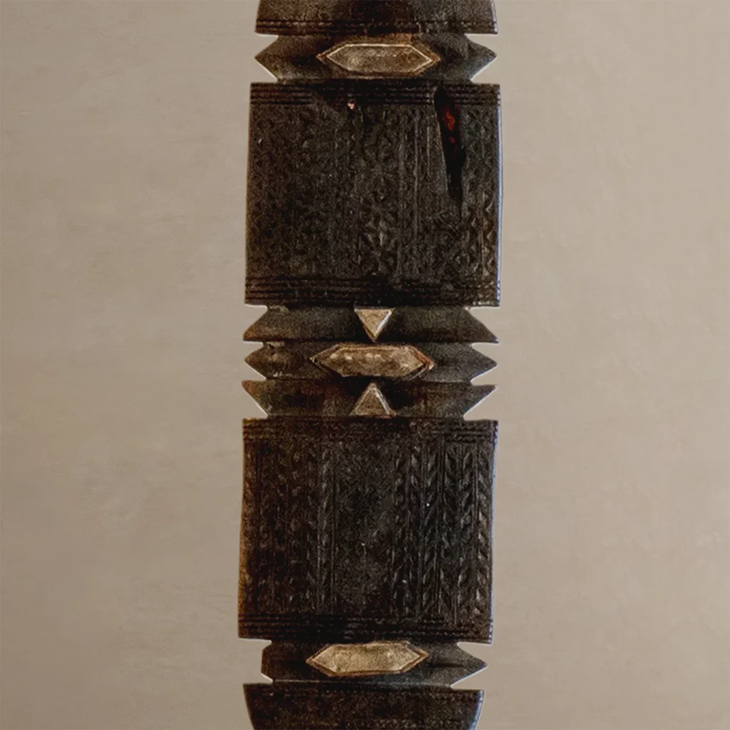 Close-up of a hand-carved, vintage wooden artifact, reminiscent of a tent peg, featuring geometric patterns and metallic embellishments on a plain background.