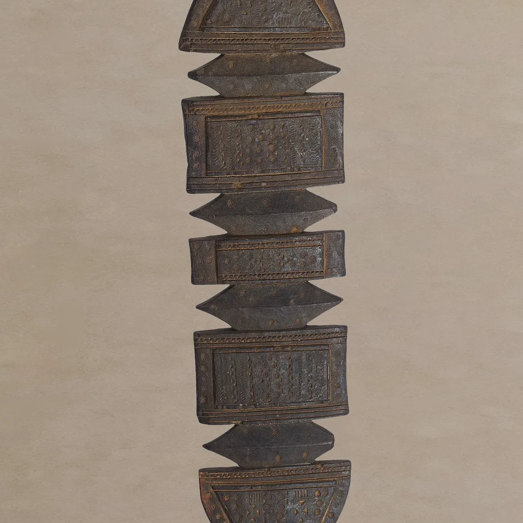 An elongated, vertical wooden sculpture with alternating square and triangular segments, featuring intricate hand-carved details reminiscent of vintage tent pegs, set against a neutral background.
