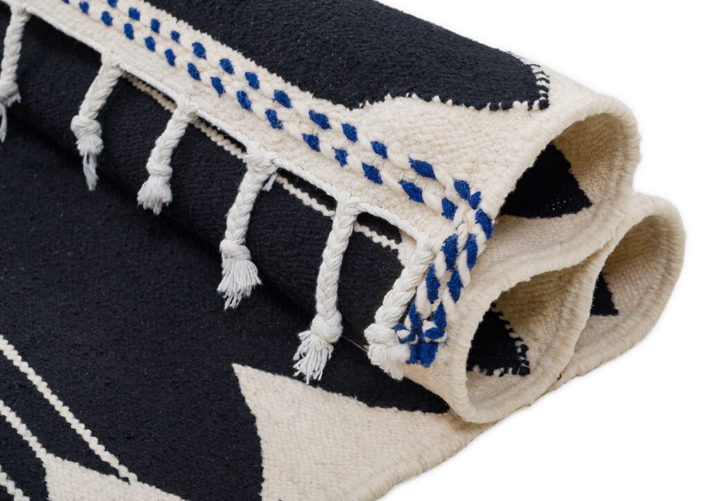 A handwoven rug in black and white, highlighted with blue accents and tasseled edges, is elegantly displayed.