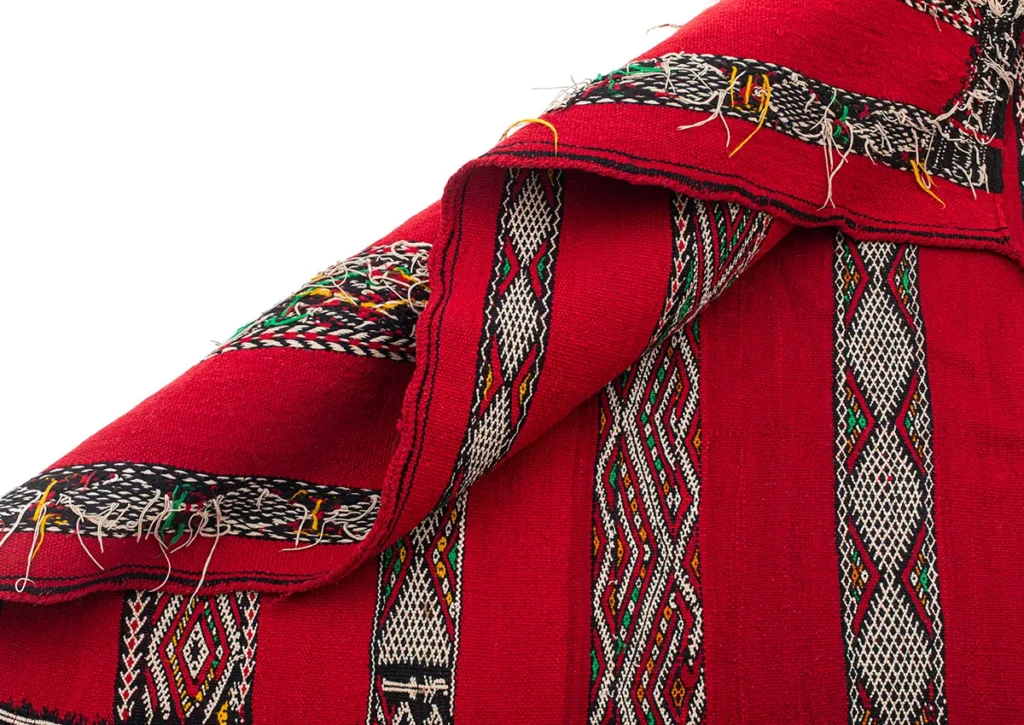 Close-up of a folded red textile, reminiscent of a vintage handwoven rug, with intricate black, white, and multicolored geometric patterns. Threads are visible along the edges.
