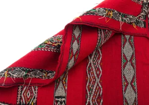 Close-up of a folded red textile, reminiscent of a vintage handwoven rug, with intricate black, white, and multicolored geometric patterns. Threads are visible along the edges.
