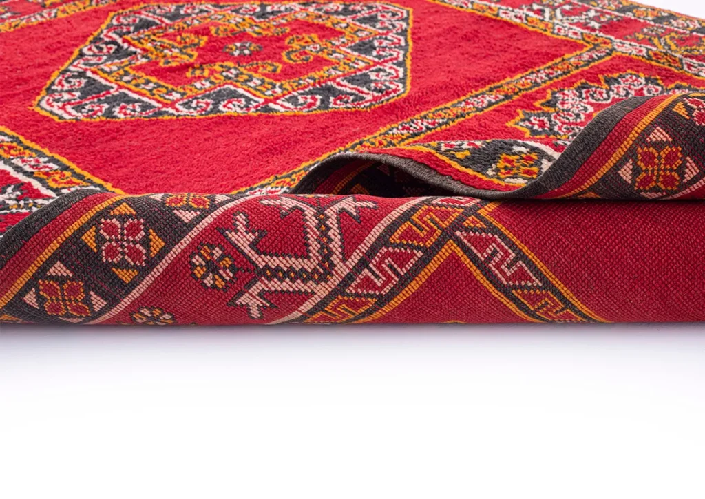 Close-up of a folded red woven rug showcasing intricate geometric patterns in black, white, and gold. The design features traditional motifs with colorful borders that highlight its rich cultural heritage.
