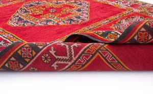 Close-up of a folded red woven rug showcasing intricate geometric patterns in black, white, and gold. The design features traditional motifs with colorful borders that highlight its rich cultural heritage.