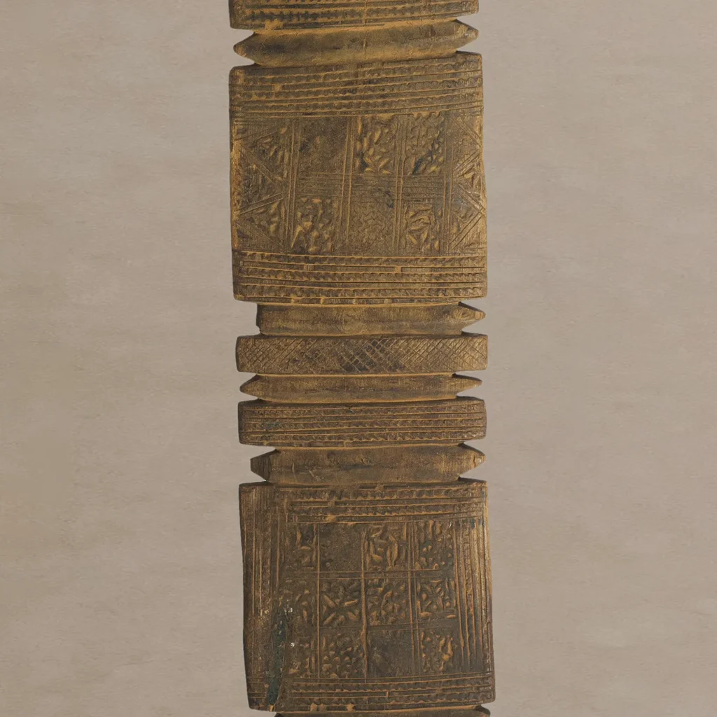 A hand-carved vintage wooden tent peg, showcasing intricate geometric patterns on a vertically aligned rectangular shape with ridges and decorative motifs on a plain background.