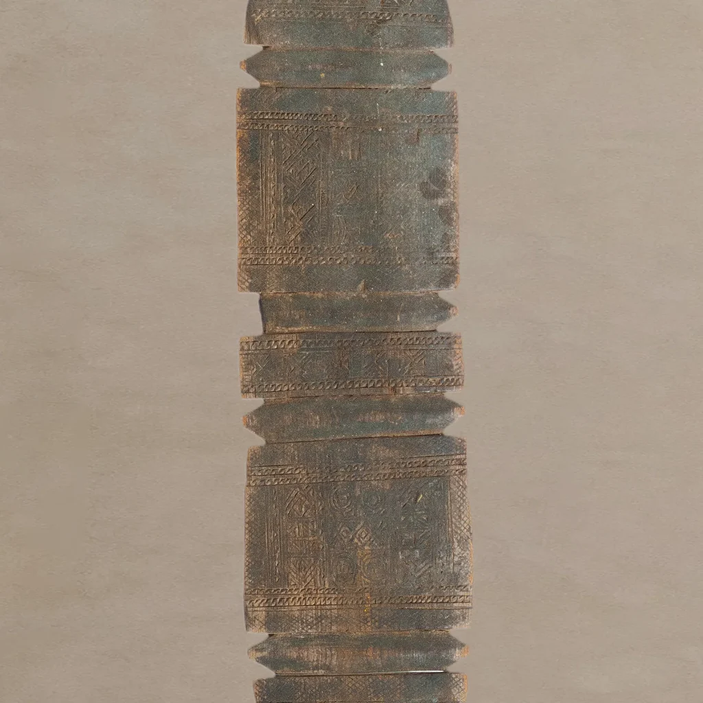 A tall, hand-carved wooden artifact with vintage geometric patterns, reminiscent of an elegant tent peg, stands vertically against a plain background.