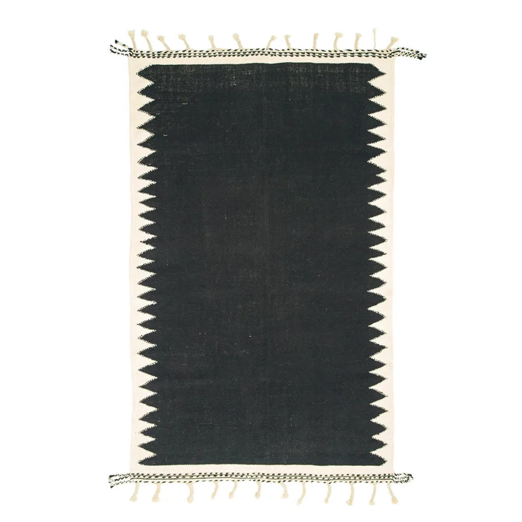 A rectangular rug features a solid dark gray center, complemented by zigzag patterned white edges. The shorter ends are accentuated with fringe details for added texture.