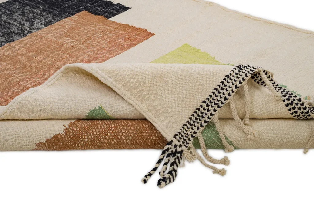 This folded woven textile features intricate geometric patterns in earthy tones, complemented by black and white fringes, creating a striking focal point on a flat surface.
