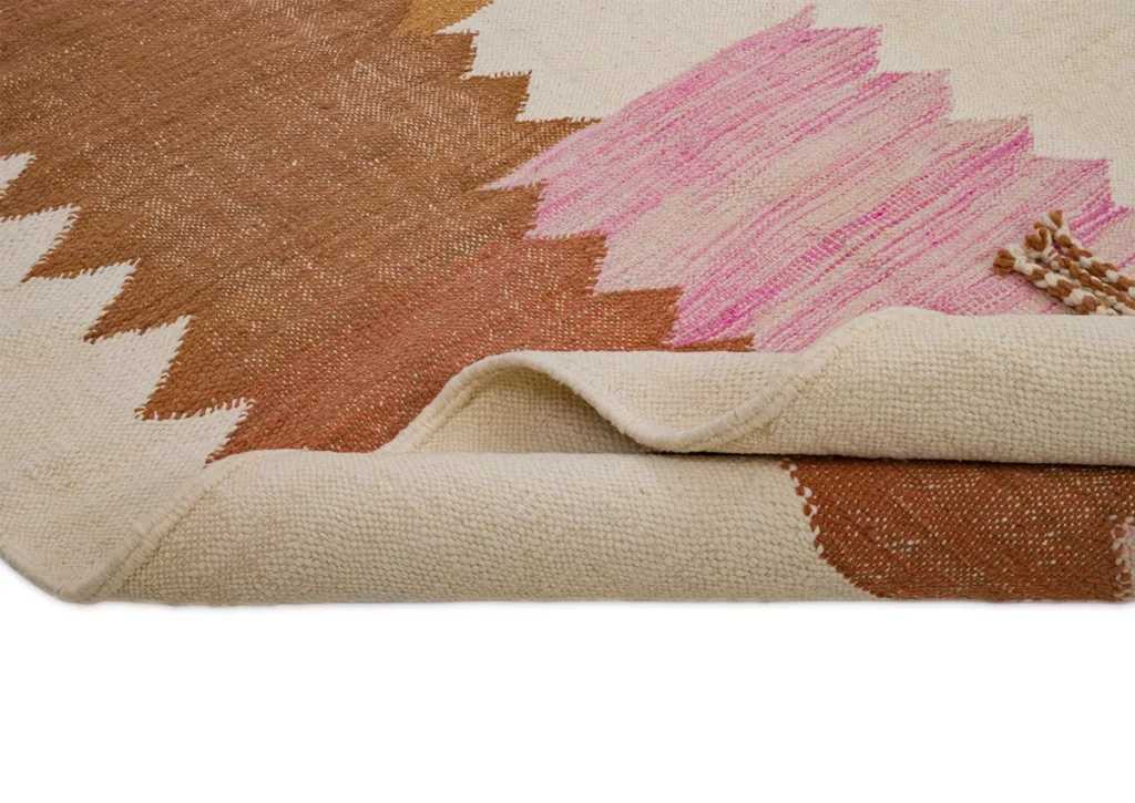 A close-up of a folded woven rug showcases a stunning geometric pattern in brown, pink, and cream colors.