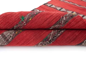 This vintage handwoven rug features a folded red textile adorned with diagonal brown and multicolored striped patterns, framed by a striking black and white geometric border.
