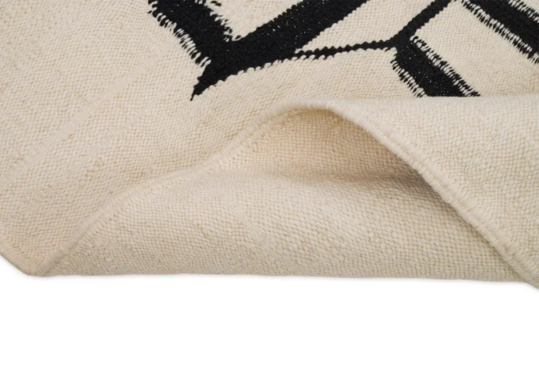 Close-up of a beige woven rug with black geometric patterns, highlighting a corner turned over to reveal the rich texture on the underside, which adds an inviting touch to your living space.