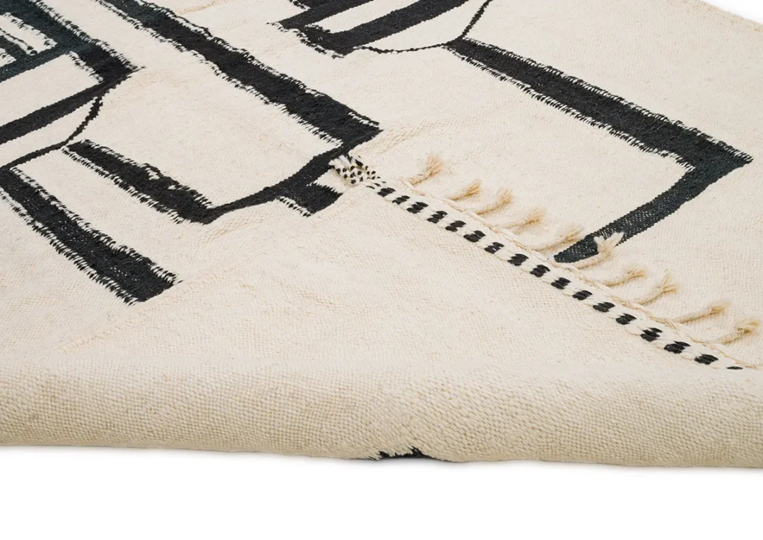 A close-up of a beige woven rug adorned with black geometric patterns and fringed edges, artistically folded.