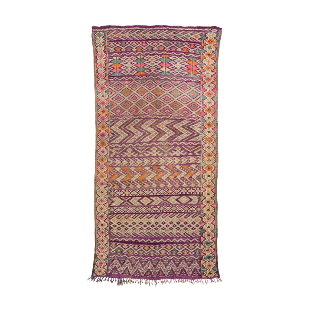 This vintage handwoven rug showcases a multicolored, intricately patterned design with geometric motifs in reds, oranges, purples, and beiges. The rugs artistry is complemented by its fringed ends.