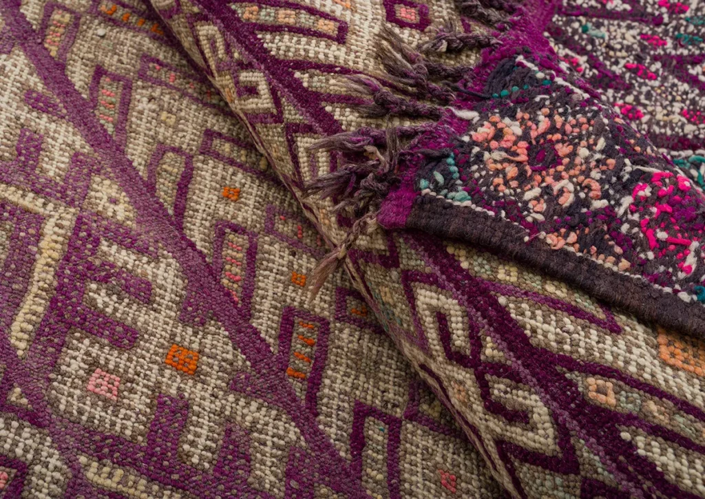 Close-up of two vintage handwoven kilim rugs showcasing intricate geometric patterns in purple, beige, and multicolored accents. Fringes delicately adorn the edge of one rug.