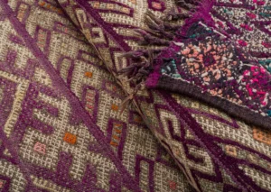 Close-up of two vintage handwoven kilim rugs showcasing intricate geometric patterns in purple, beige, and multicolored accents. Fringes delicately adorn the edge of one rug.