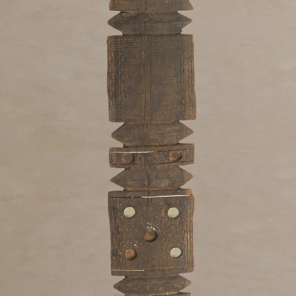 A carved wooden staff featuring vintage geometric patterns, reminiscent of the intricate designs found on traditional tent pegs, adorned with circular metal embellishments.