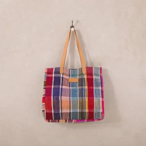 A multicolored plaid tote bag with tan leather handles hangs on a wall hook against a light beige background.