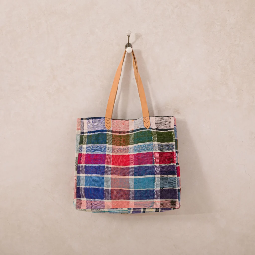 A colorful plaid tote bag with tan leather handles hangs on a wall hook.