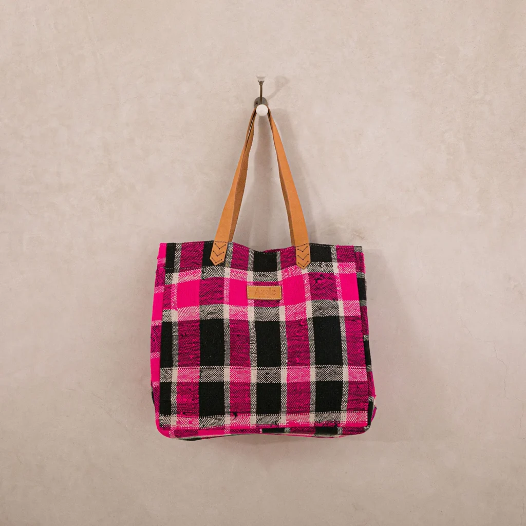 An upcycled shopper bag in pink and black plaid with brown handles hangs on a metal hook against a plain beige wall.