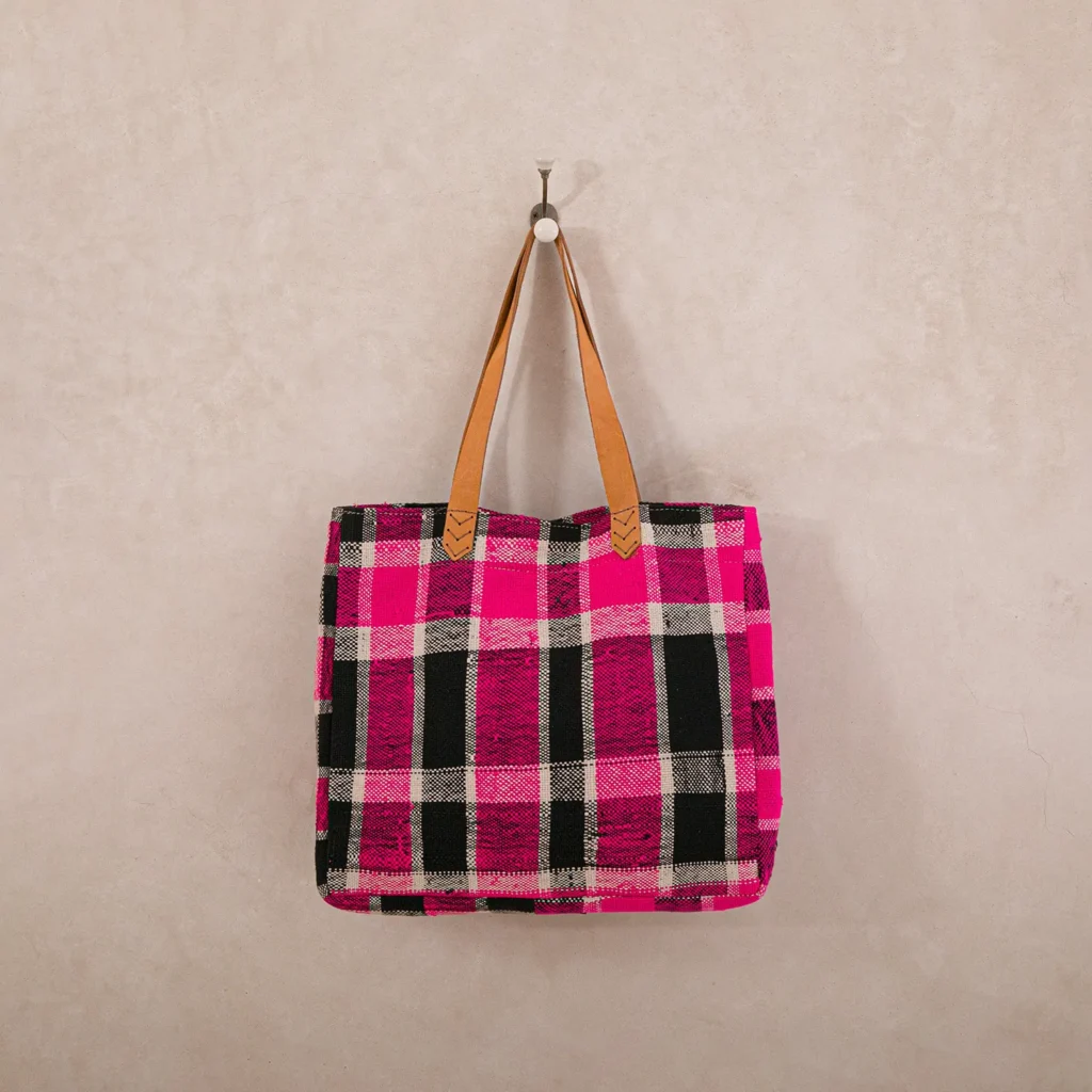An upcycled shopper bag in pink, black, and white plaid with brown handles hangs stylishly on a hook against a beige wall.