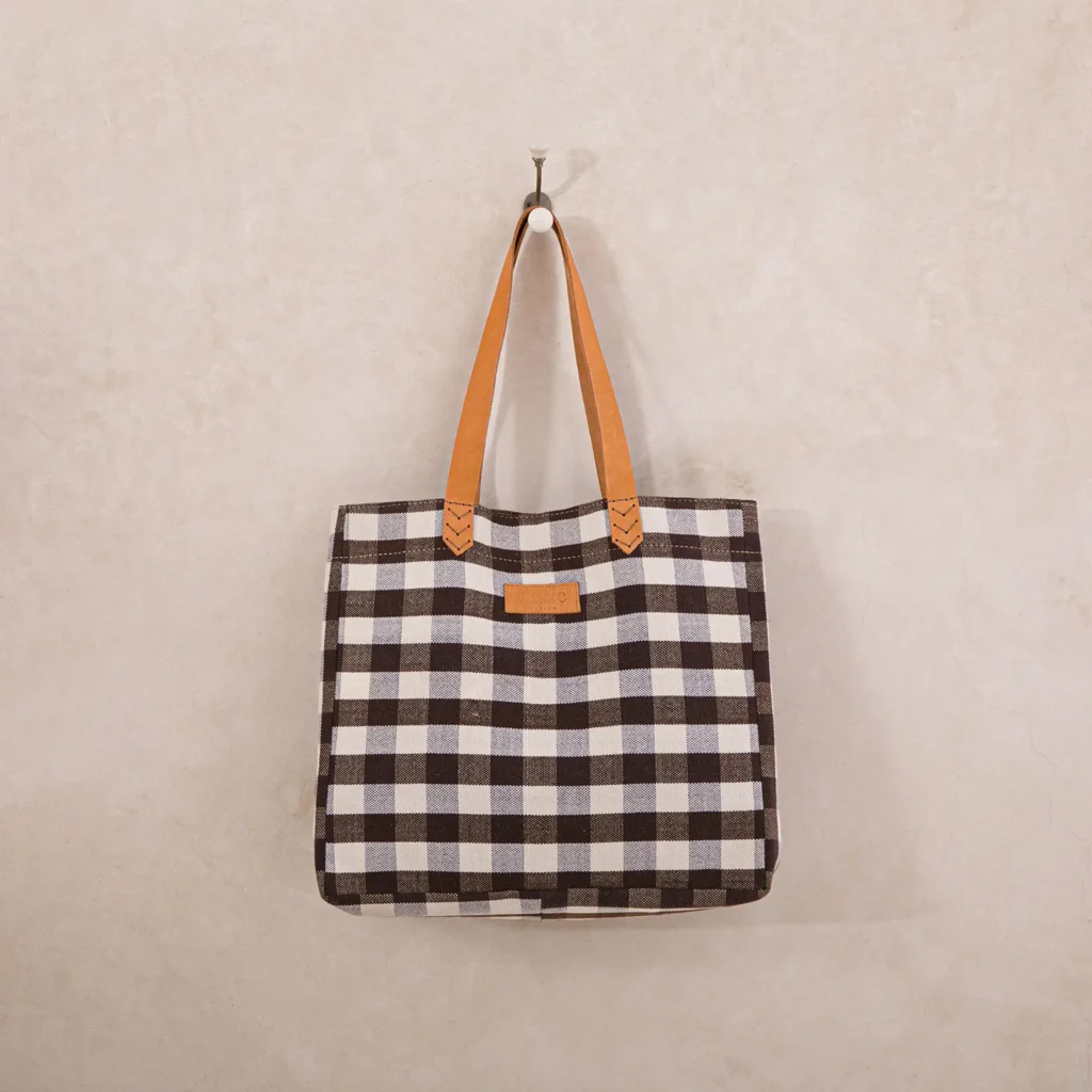 A black and white checkered tote bag with tan leather handles hangs on a single hook against a light beige wall.