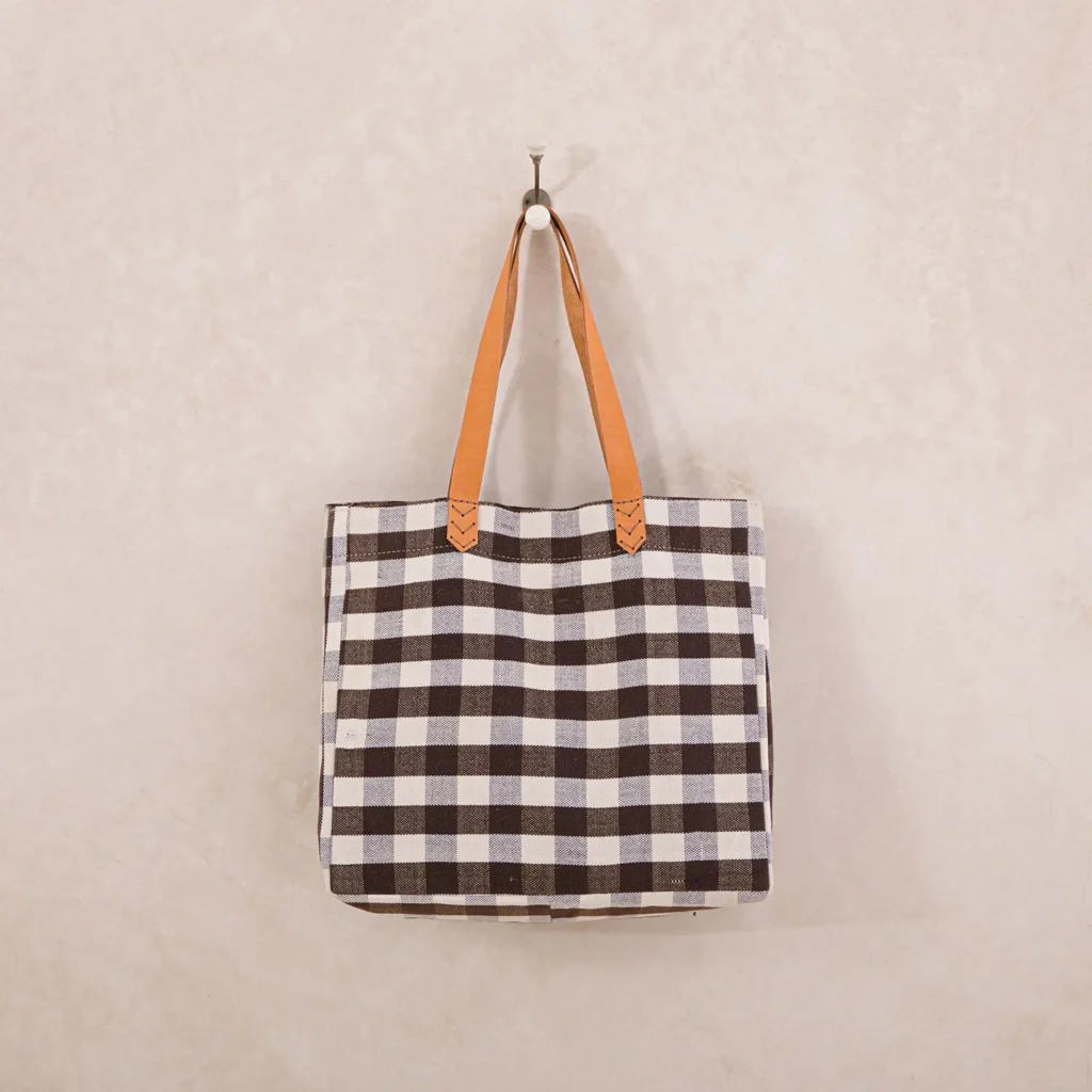 A checkered tote bag with brown leather handles hangs on a wall.