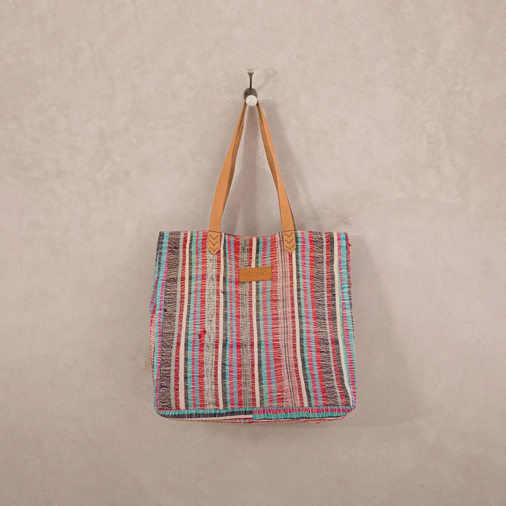 A multicolored striped tote bag with brown leather handles hangs on a white hook against a beige wall.