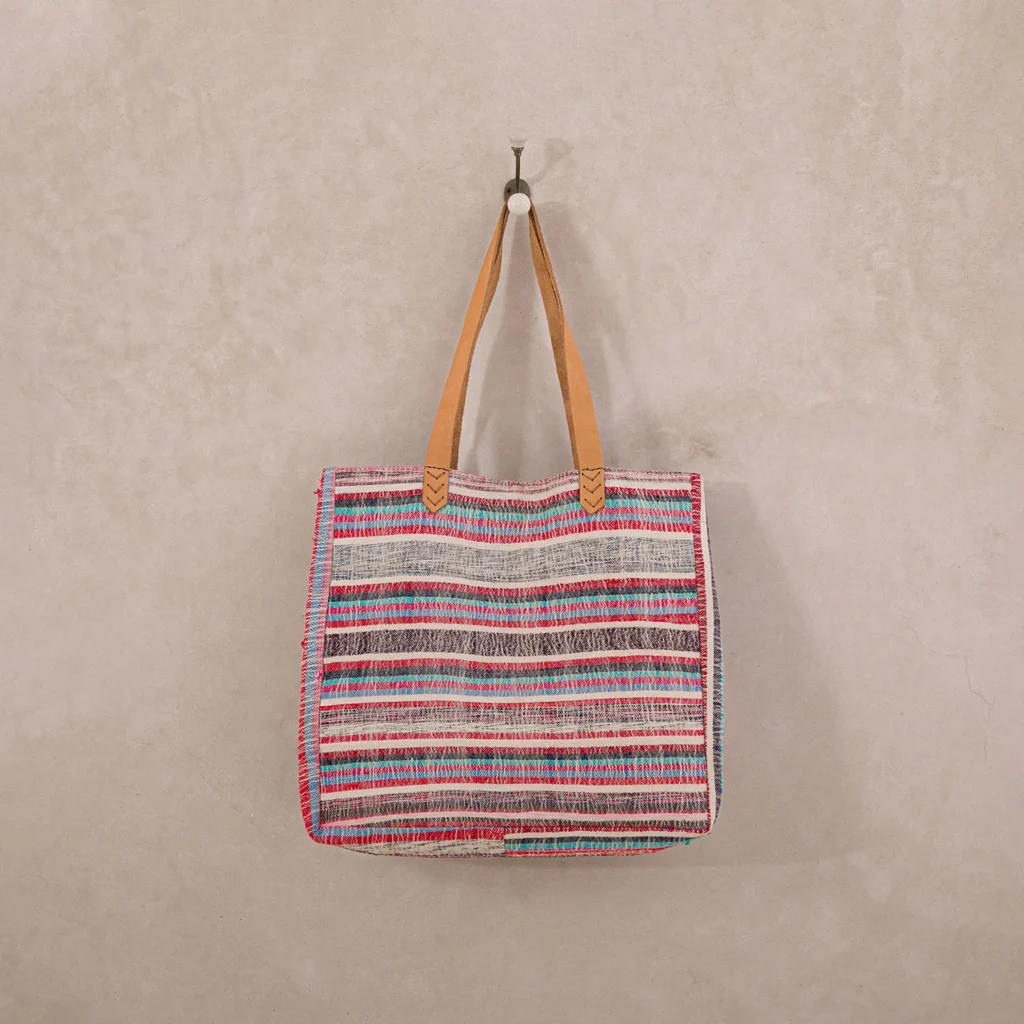 Colorful woven tote bag with tan leather handles hangs on a wall hook against a beige background.