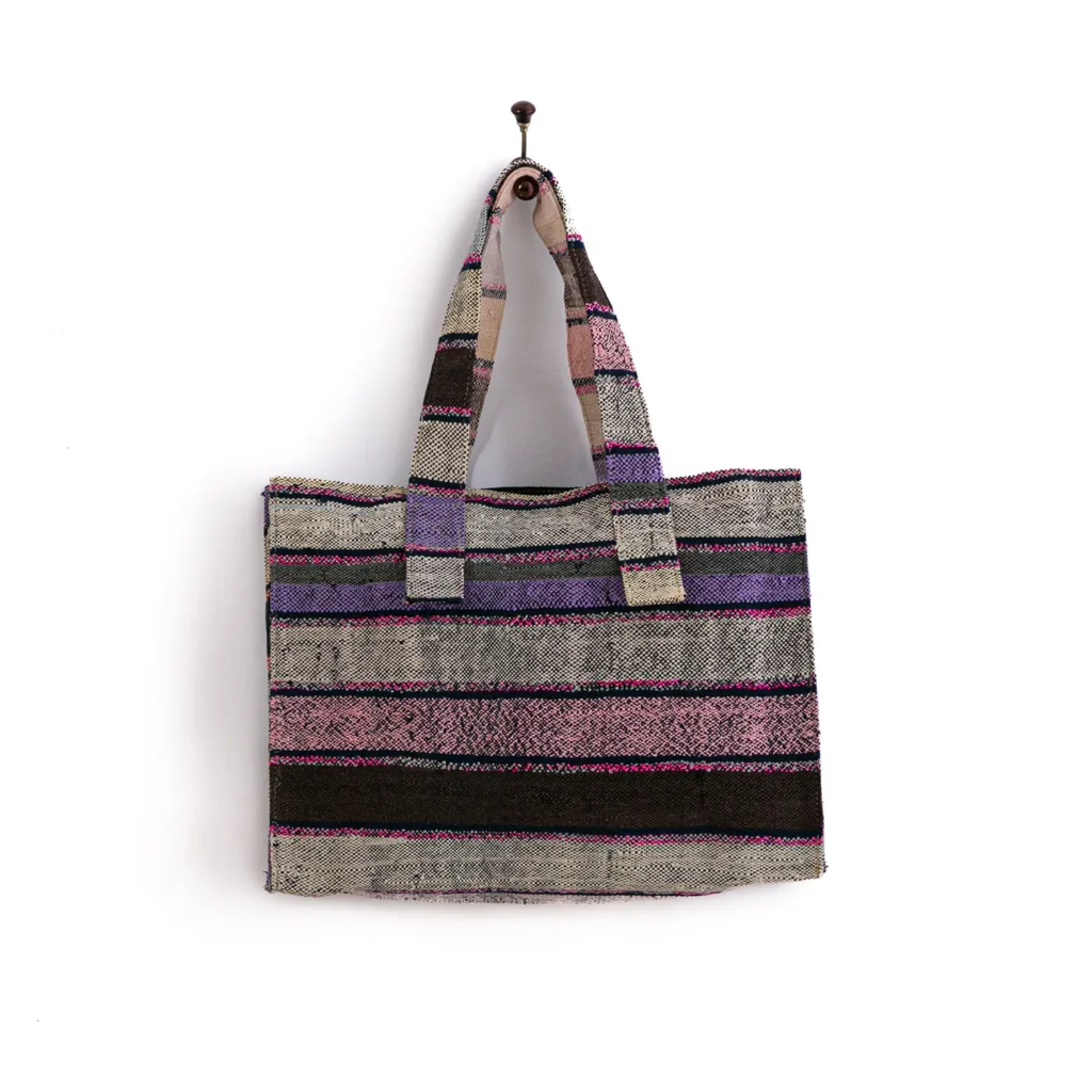 A multicolored striped woven tote bag hanging on a single hook against a plain white background. The bag features shades of purple, black, and beige.