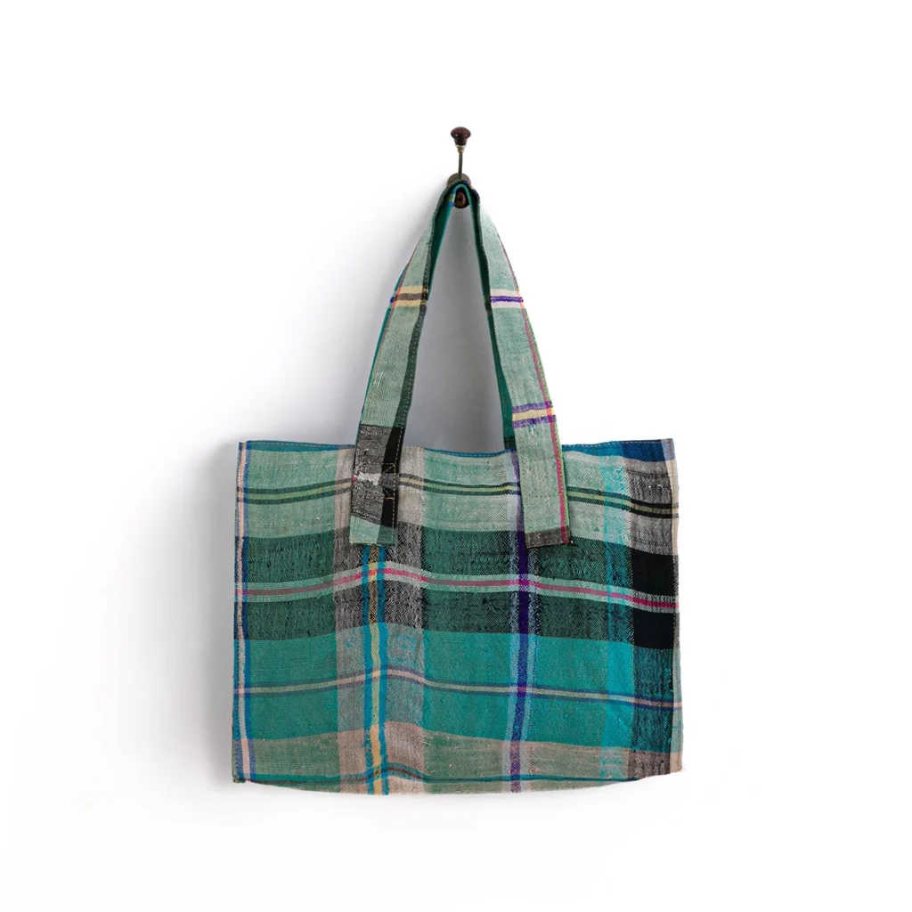 A plaid pattern tote bag with shades of green, blue, pink, and black hangs on a simple wall hook against a white background.