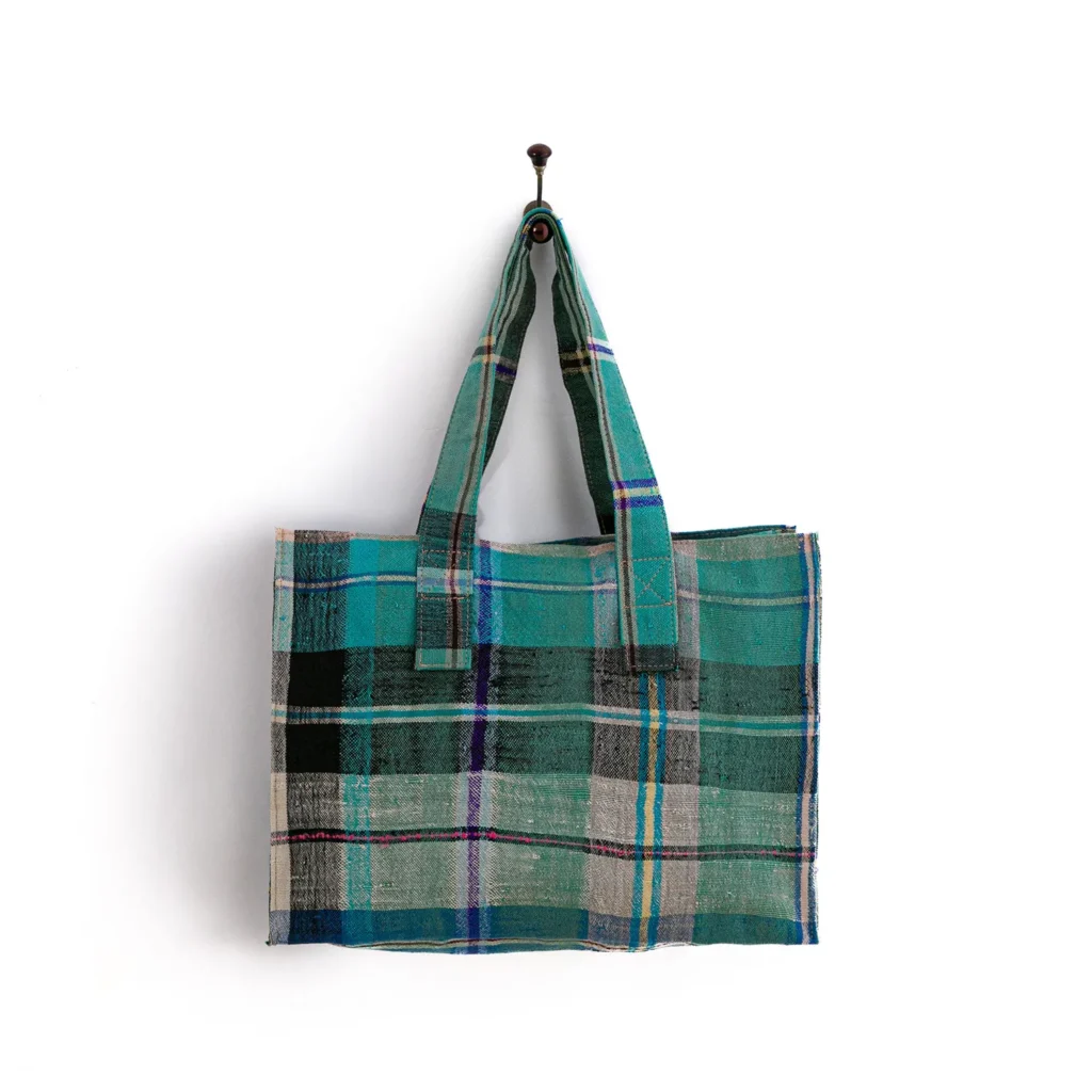A plaid tote bag with shades of green, blue, and beige hangs from a hook on a white wall.